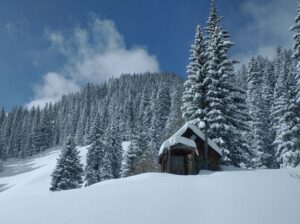 deep snow covers a small house and pine trees below a blue sky with wispy clouds