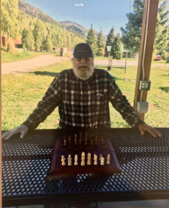 a bearded man at a shaded table in the park, sitting in front of a chess board, invites you to make the first move